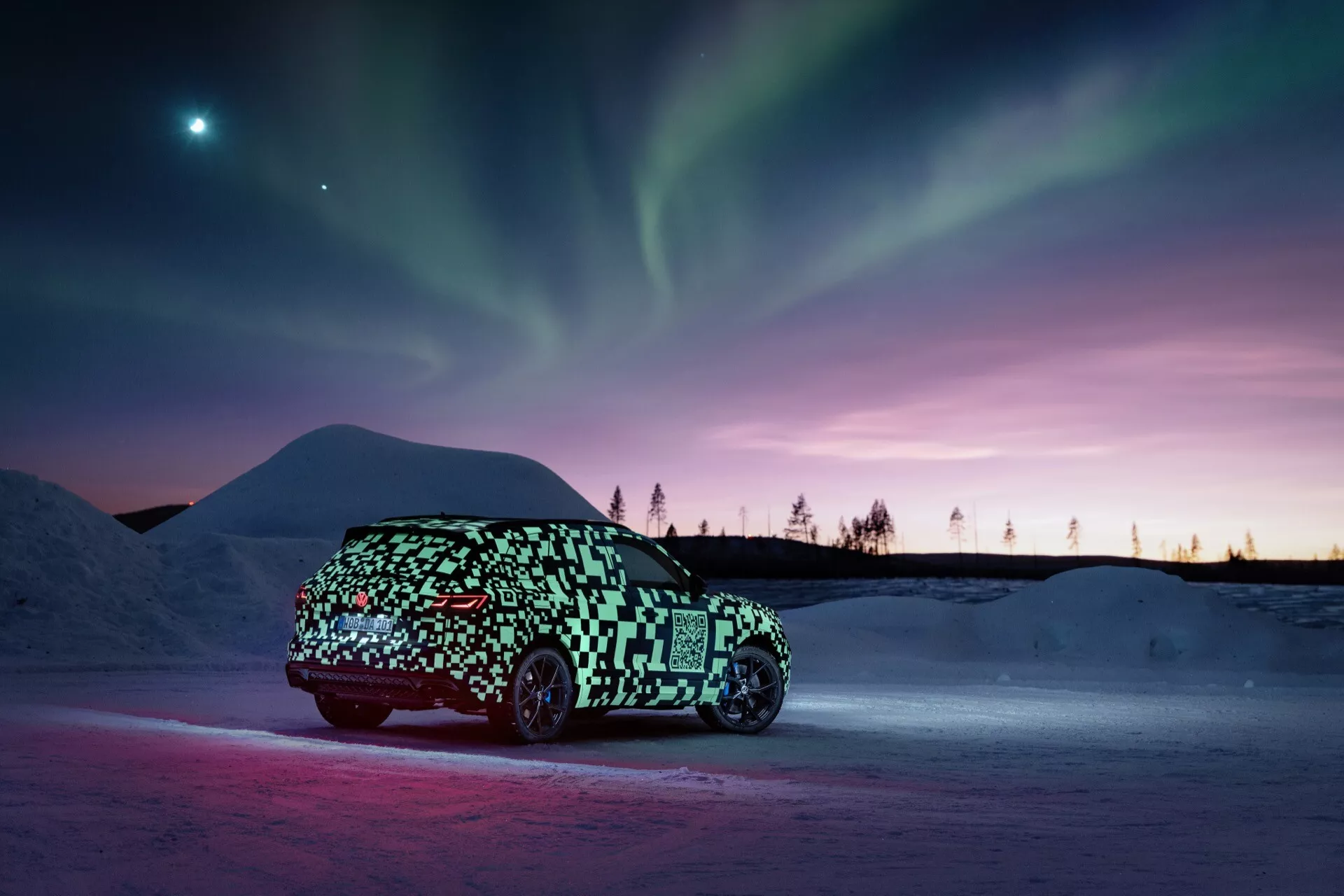 Prototyp VW Touareg (III-Facelift) 2023 w kamuflażu uchwycony z profilu bocznego na śnieżnym podłożu z ambientowym oświetleniem podkreślającym pojazd na tle zorzy polarnej i odcieni zmierzchu, sugerujący elegancki, możliwie elektryczny, design hatchbacka lub crossovera