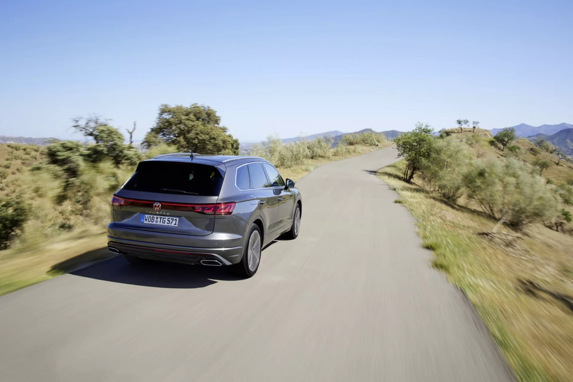 Widok z tyłu trzy czwarte VW Touareg III-Facelift 2023 na malowniczej drodze, ukazujący LED-owe światła tylne, charakterystyczne oznaczenia, podwójne końcówki wydechu, aluminiowe koła, elegancką linię dachu i błyszczące wykończenie.