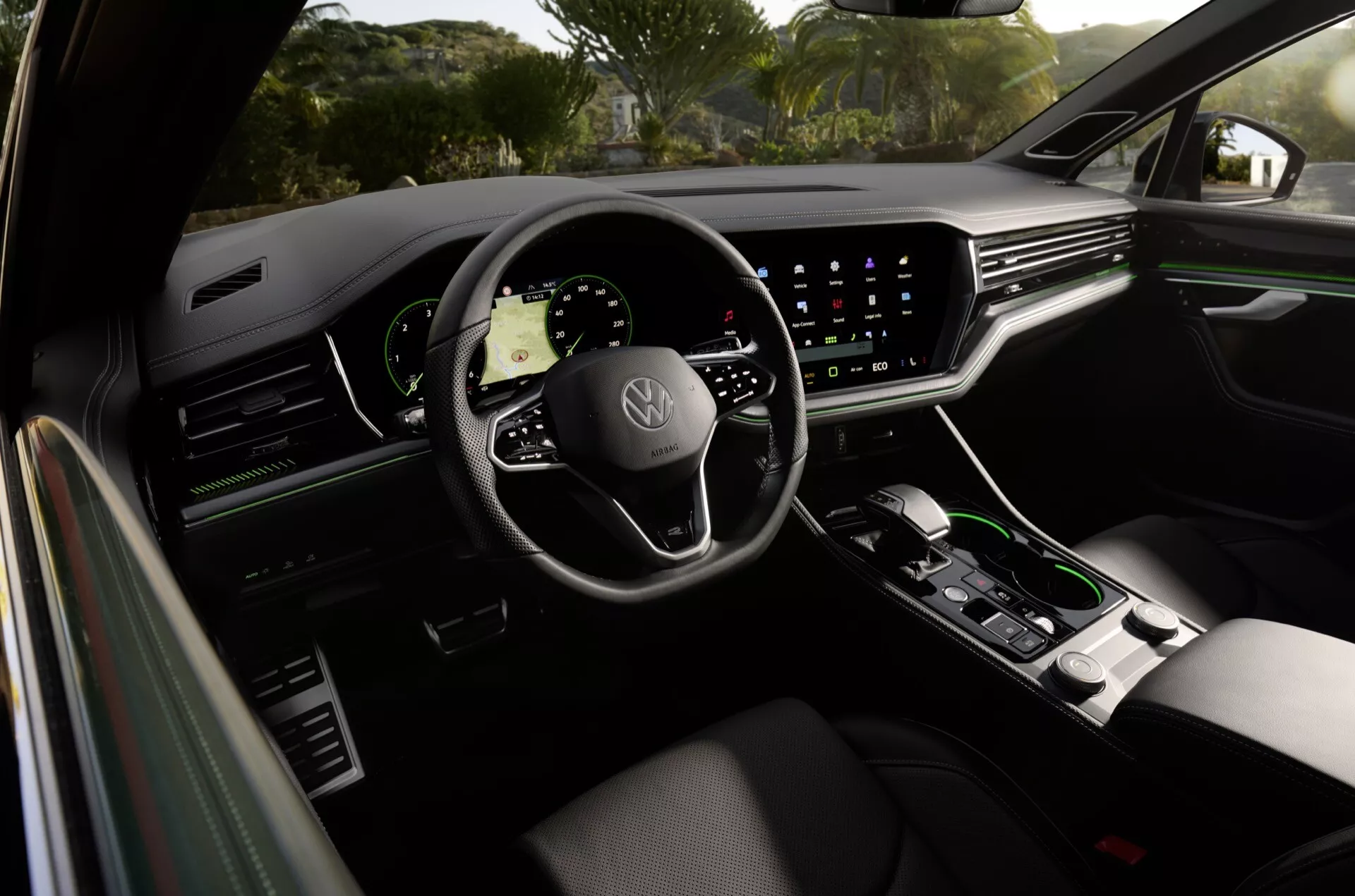 Wnętrze Volkswagen Touareg 2023 widziane z tylnego siedzenia, przedstawiające panel drzwi kierowcy, premium ergonomiczne siedzenie kierowcy, kierownica z logo Volkswagen z zamontowanymi sterowaniami, cyfrowy panel wskaźników i duży system infotainment z ekranem dotykowym z wyświetlaną nawigacją, a także minimalistyczne sterowanie klimatyzacją i selektor automatycznej skrzyni biegów na konsoli środkowej, odzwierciedlające luksusowe cechy i zaawansowaną technologię