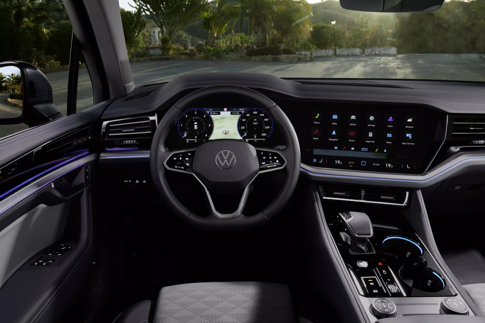 Widok wnętrza VW Touareg 2023 z perspektywy kierowcy, pokazujący nowoczesną kierownicę z trzema ramionami z logo Volkswagen, cyfrowy panel wskaźników, szeroki ekran dotykowy systemu infotainment, oświetlenie ambientowe, elegancką konsolę środkową z selektorem biegów i materiały premium