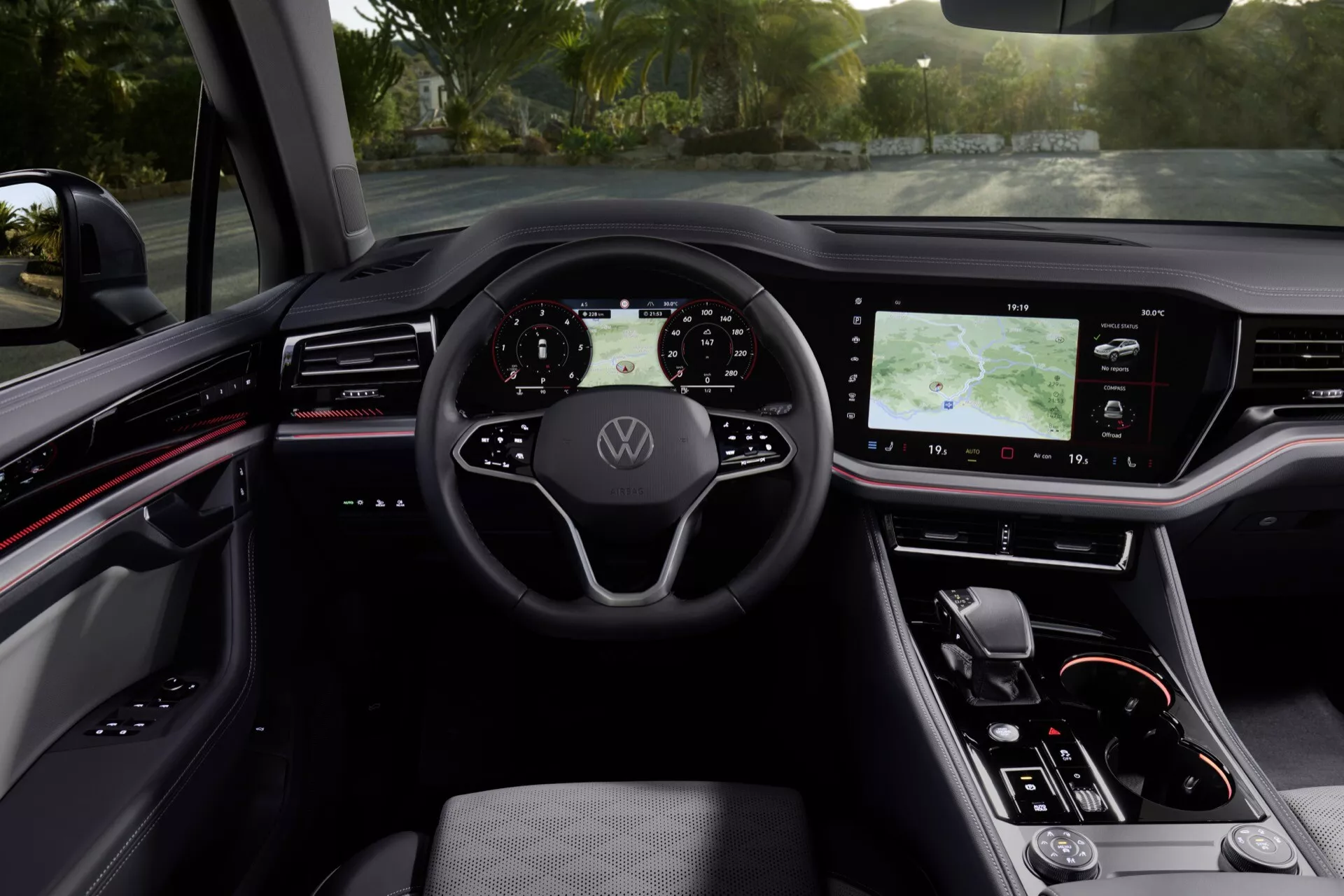 Wnętrze VW Touareg 2023 prezentujące kierownicę z logo Volkswagen, cyfrowy kokpit, duży system informacyjno-rozrywkowy na konsoli środkowej, ergonomiczne siedzenie kierowcy z materiału przypominającego skórę i nowoczesne elementy designu z czerwonymi akcentami z perspektywy kierowcy.