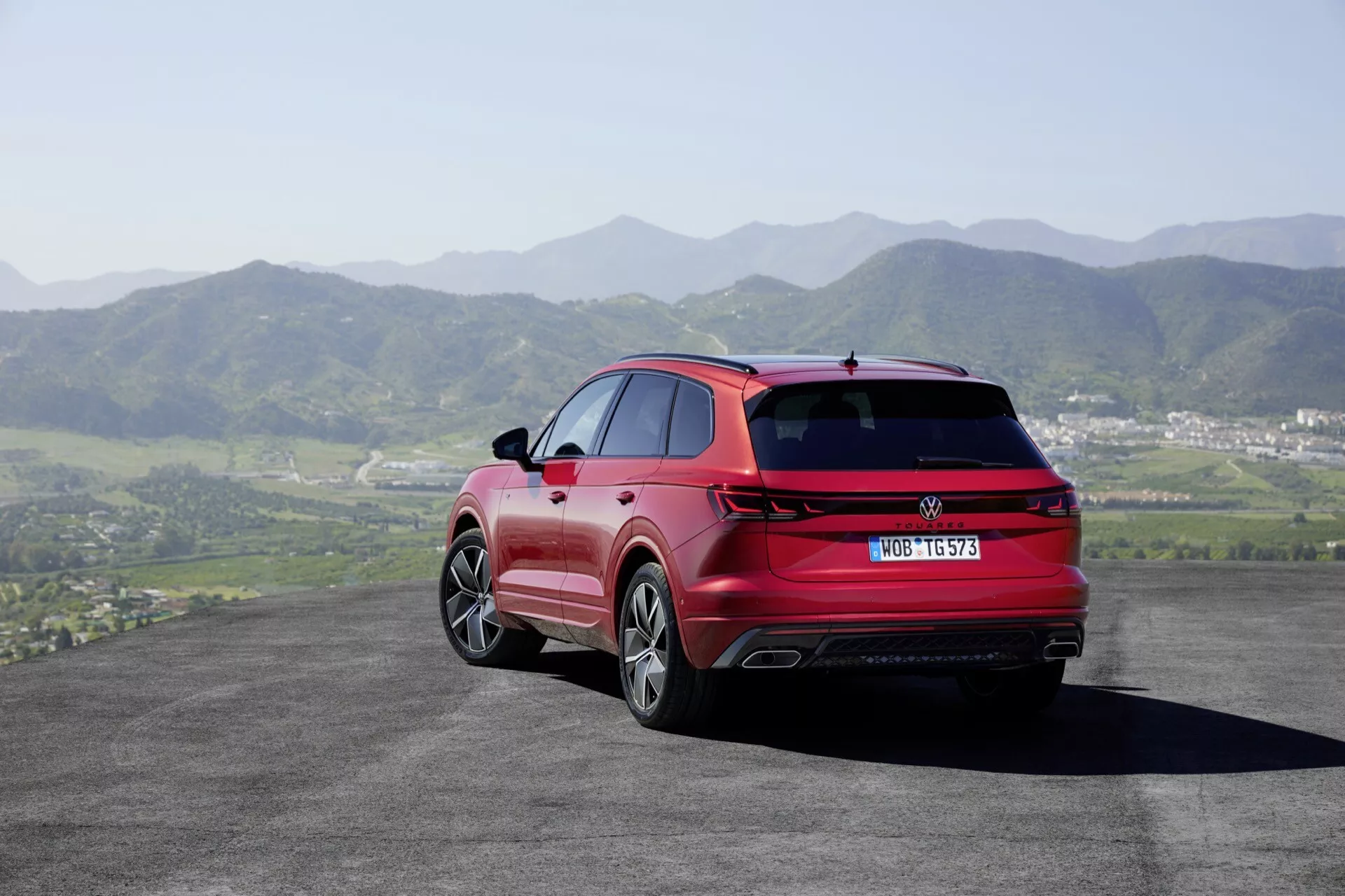 VW Touareg III Facelift 2023 w kolorze czerwonym - widok z tylu i boku ukazujący diody LED w tylnych światłach, podwójne końcówki wydechu, alufelgi i srebrne relingi dachowe na tle pagórkowatego krajobrazu i czystego nieba