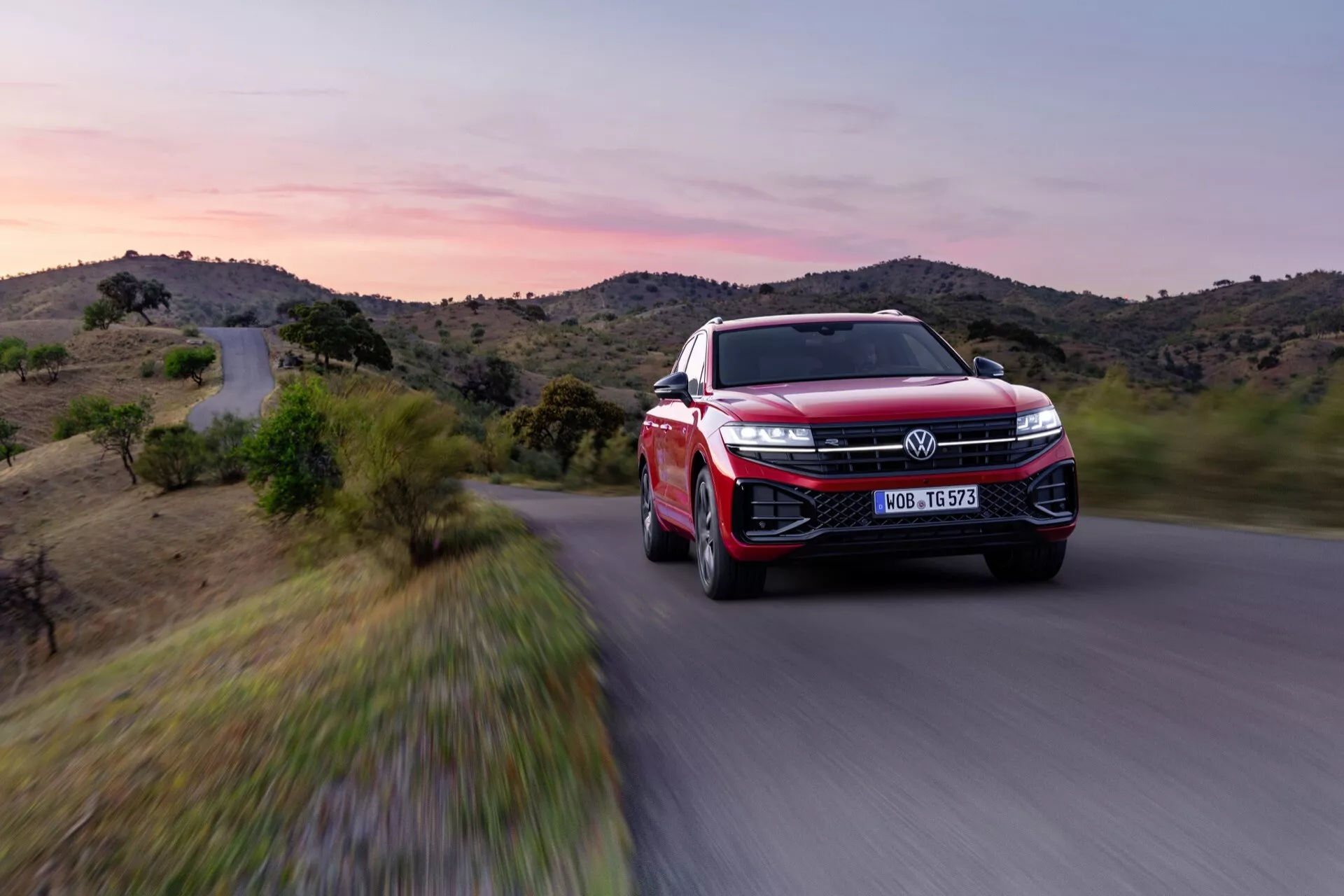 VW Touareg III-facelift 2023 w kolorze czerwonym, pokazujący widok z przodu pod kątem trzech czwartych z akcentem na reflektory LED, dużą kratkę z emblematem, sportowy design aerodynamiczny, zaawansowane koła z aluminiowych stopów i funkcje ADAS w dynamicznym ruchu na malowniczej drodze o zmierzchu