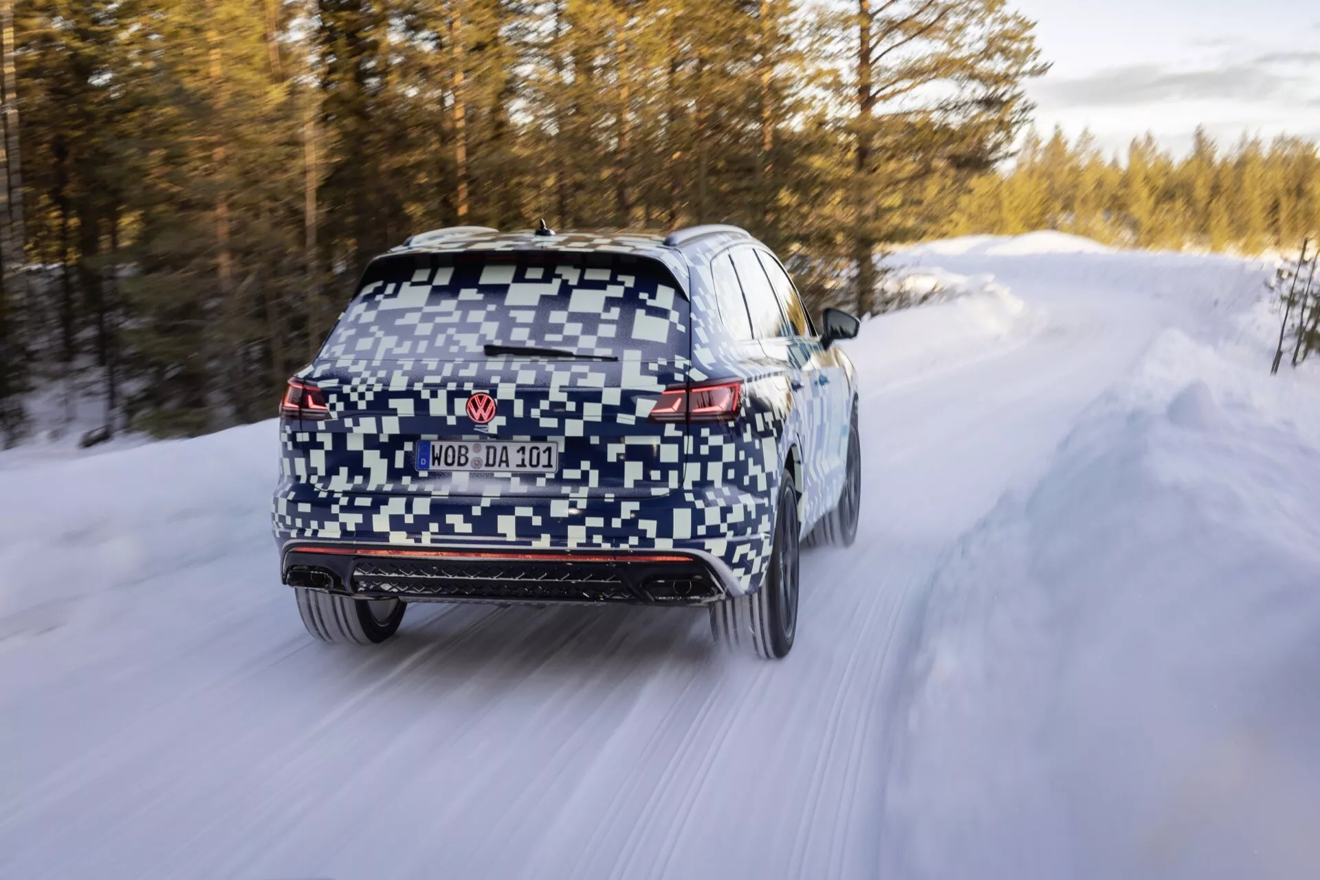 VW Touareg III Facelift 2023 w kamuflażu jadący po zaśnieżonej drodze, z LED-owymi światłami tylnymi, tylnym dyfuzorem, wycieraczką tylnej szyby, częściowo ukrytą tablicą rejestracyjną, podwójnymi końcówkami wydechu, czujnikami na tylnym zderzaku, rozmyciem ruchu wskazującym ruch, oponami zimowymi, i potencjalnym napędem na wszystkie koła