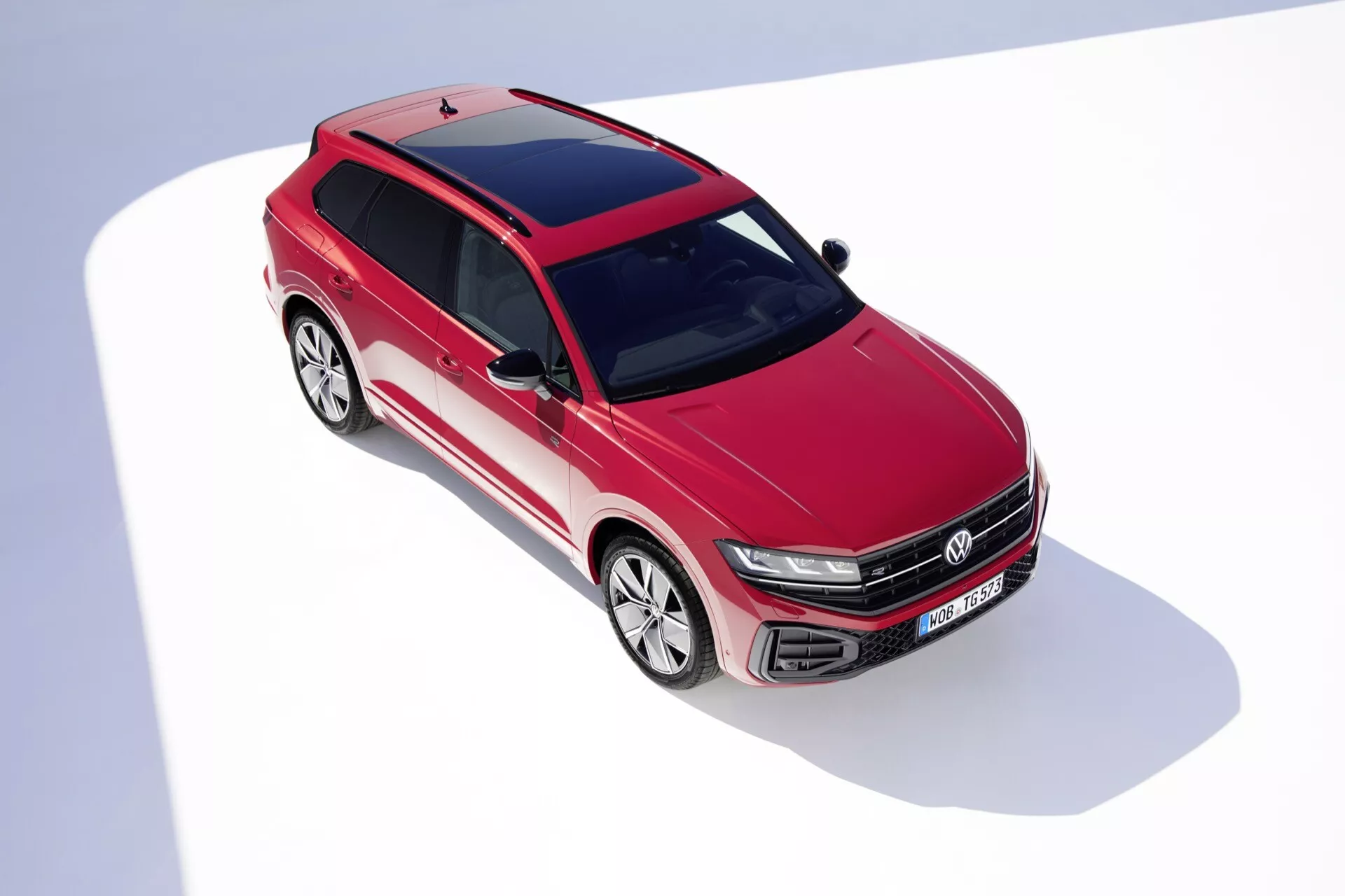Widok z wysokiego kąta przedniej części VW Touareg III Facelift 2023 w kolorze czerwonym, prezentujący cechy konstrukcyjne, w tym grill z poziomymi listwami i emblematem, reflektory LED, aerodynamicznie stylizowany przedni zderzak z wlotami powietrza, rzeźbione linie maski, obszerne przednie szyby, koła z lekkich stopów o wieloramiennym wzorze, panoramiczny dach oraz aerodynamiczne lusterka boczne na białym tle