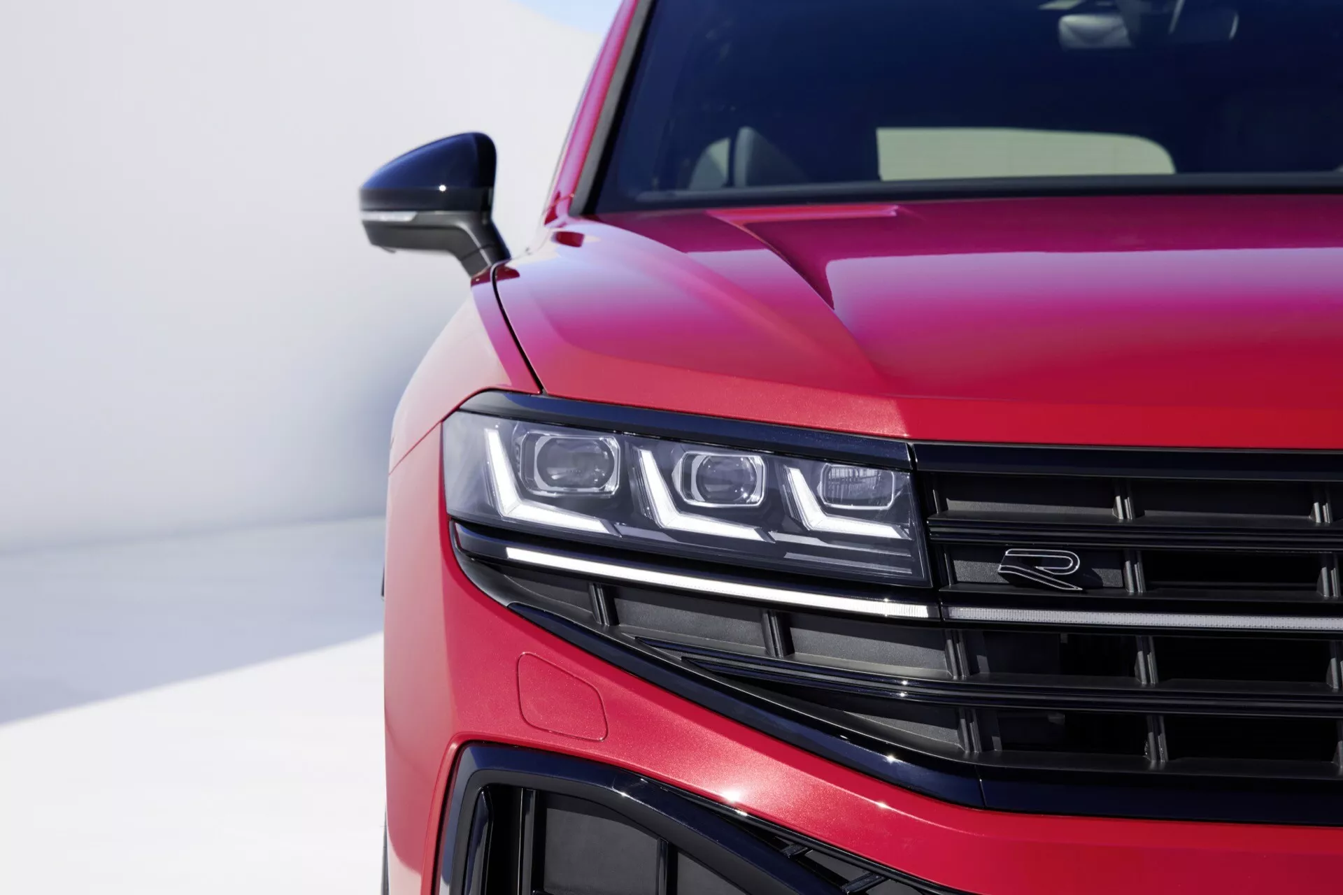 VW Touareg III-Facelift 2023 w lśniącym metalicznym czerwonym lakierze z bliskim widokiem na przedni lewy kwartał, pokazujący zespół reflektorów LED, część grilla z poziomymi listwami, szczegółowy widok przedniego zderzaka i aerodynamiczne linie na masce, na rozmytym tle podkreślającym nowoczesny design i cechy technologiczne samochodu.