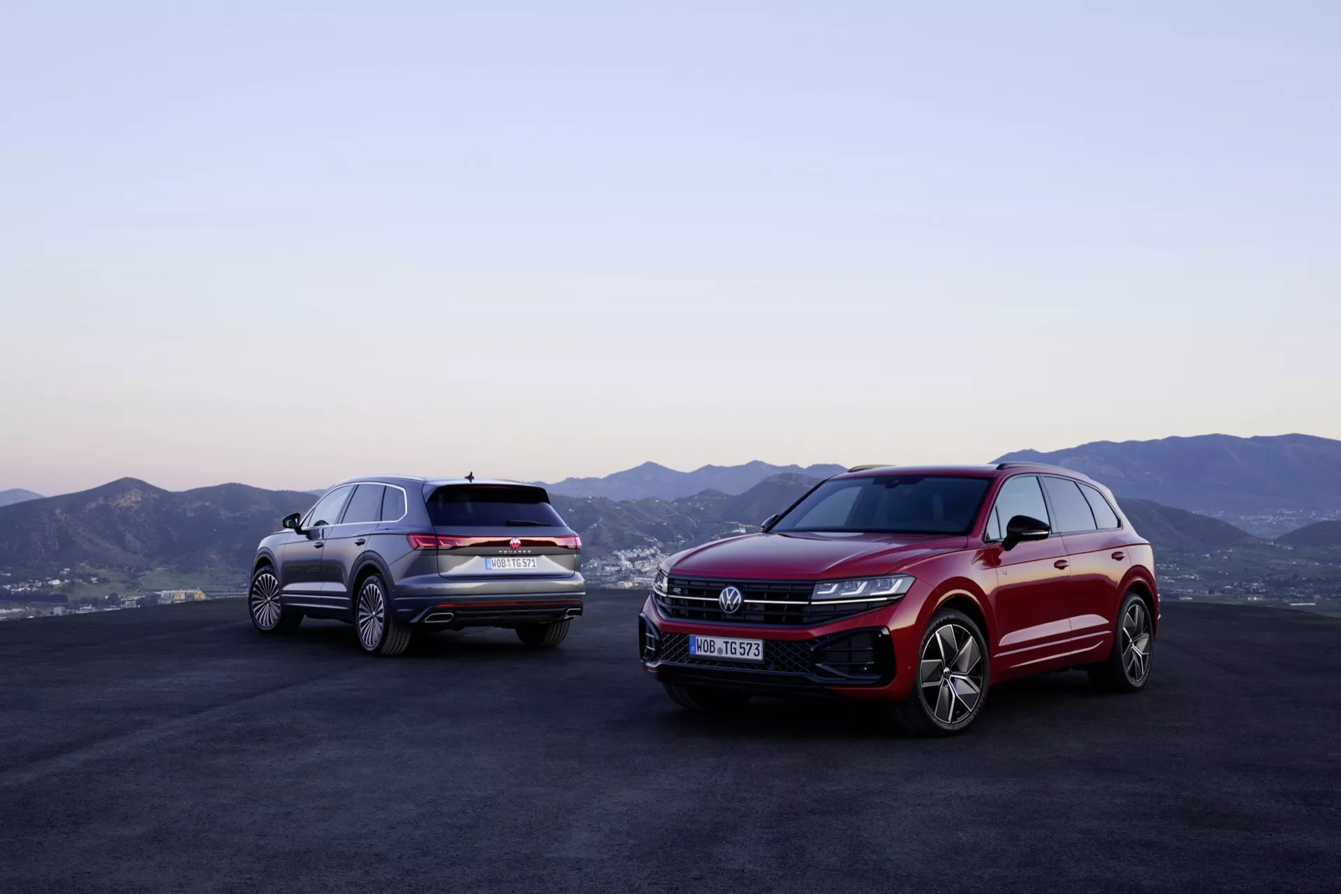 Modele Volkswagen Touareg (III-facelift) 2023 w kolorach czerwonym i srebrnym na zewnątrz z górskim pejzażem w tle i zmierzchowym niebem, prezentujące profile przednie i boczne z zaawansowaną technologią oświetlenia LED