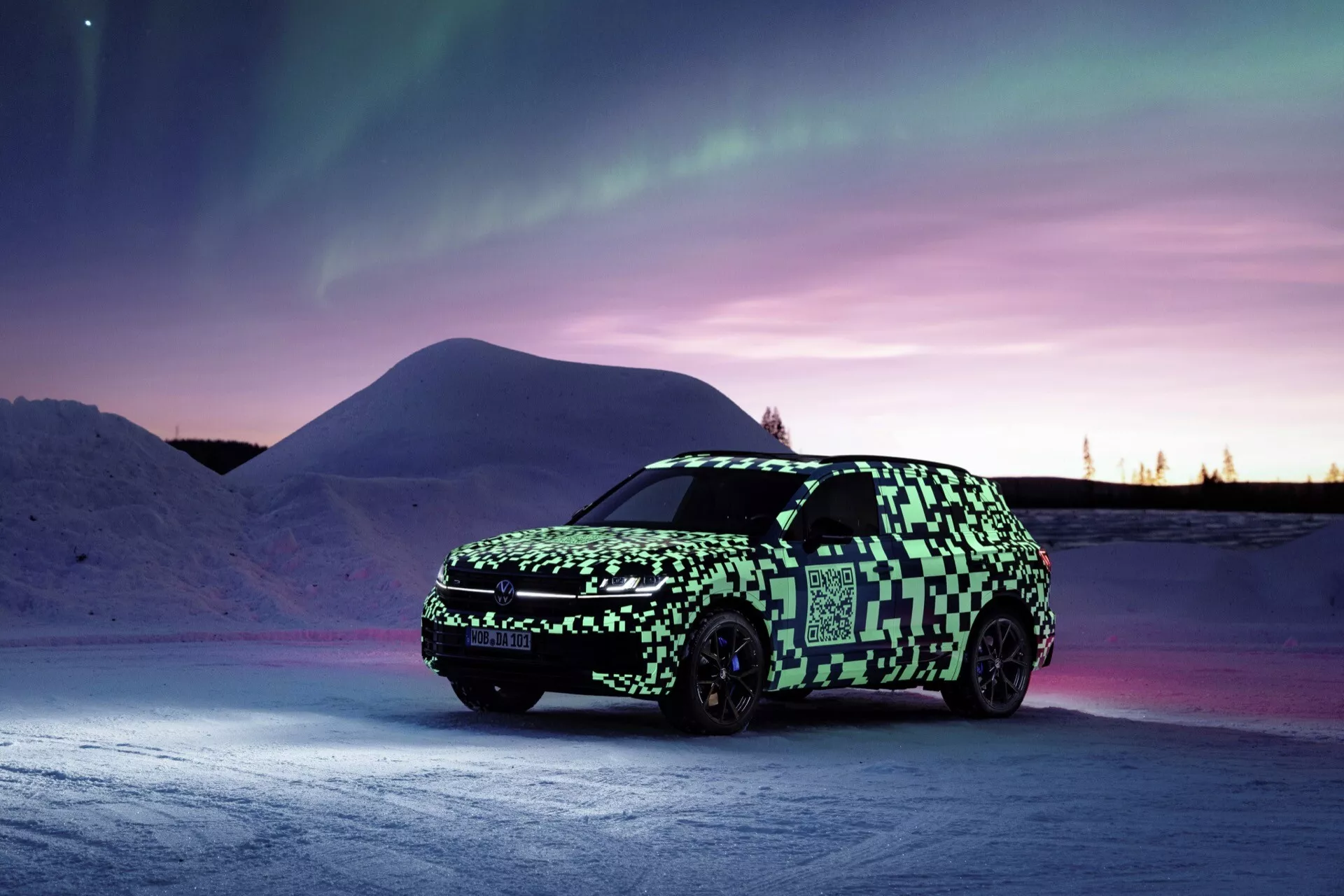 Model Volkswagen Touareg III facelift 2023 prezentowany w zimowym zmierzchu, widok z przodu pod kątem trzech czwartych z naciskiem na reflektory LED, grill przedni i kamuflaż na tle zorzy polarnej i śnieżnego krajobrazu