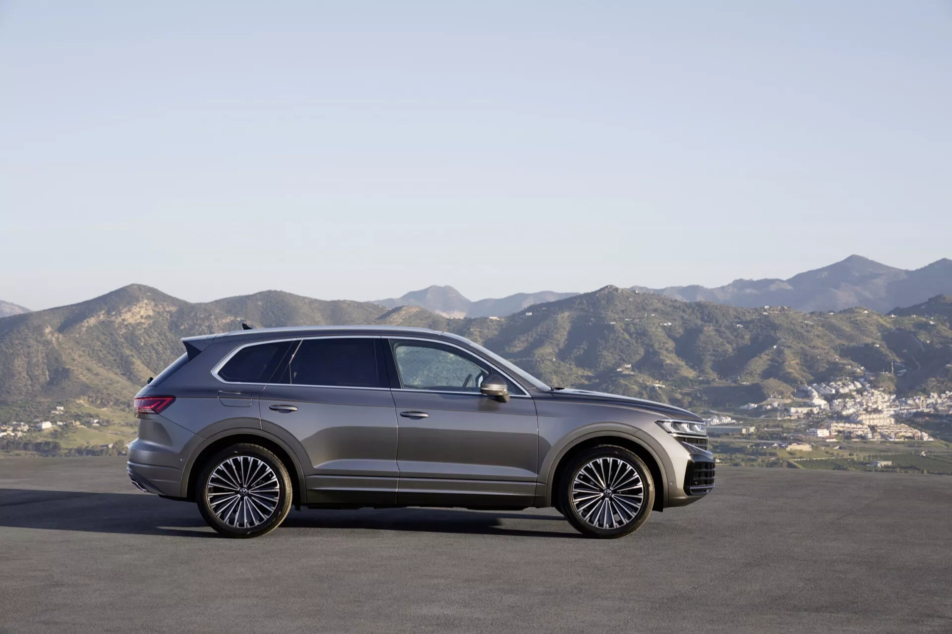 VW Touareg III po liftingu z 2023 roku w widoku z boku zaparkowany na płaskiej powierzchni na tle górskiego krajobrazu pod jasnym niebem, prezentujący jego nowoczesny design SUV, stylowe felgi aluminiowe i panoramiczny dach.