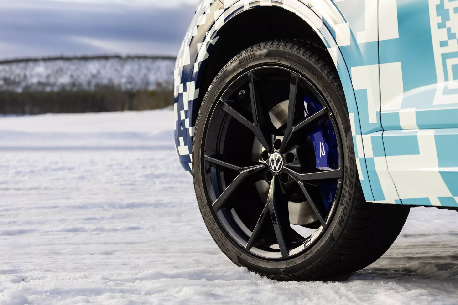 Zbliżenie na przednie koło VW Touareg III Facelift 2023 z czarną felgą wielowiązkową, logo Volkswagen i niebieskimi zaciskami hamulcowymi na śnieżnej powierzchni, prezentujące funkcje wydajnościowe i testy zimowe