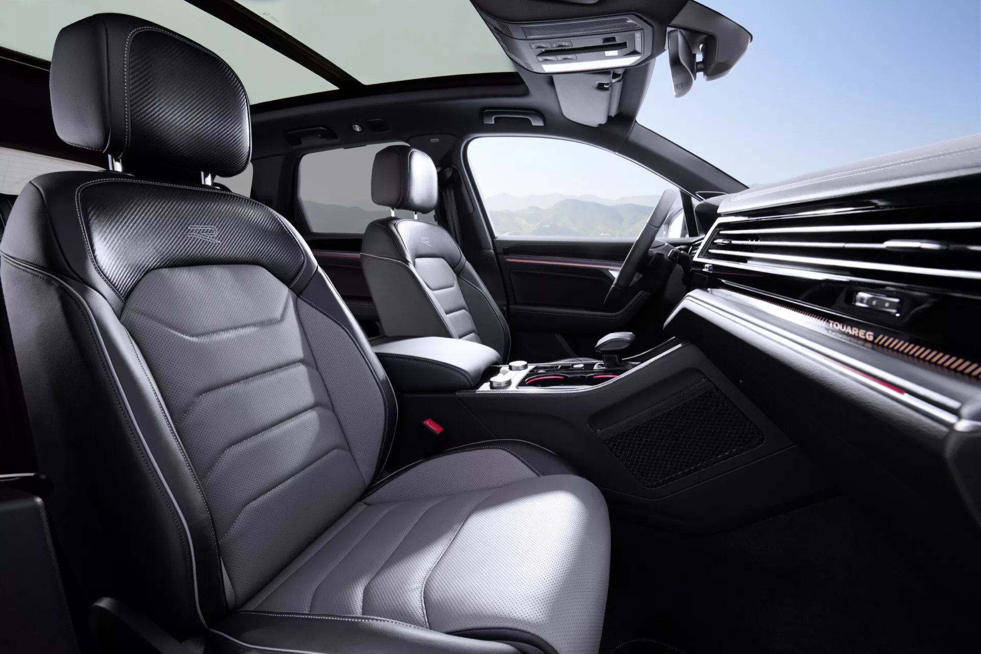 Wnętrze VW Touareg III Facelift 2023 z perspektywy tylnego siedzenia, pokazujące przedni obszar pasażera i kierowcy, z luksusowymi siedzeniami tapicerowanymi z ozdobnymi szwami, obszerną deską rozdzielczą z eleganckimi wylotami powietrza i centralnym ekranem multimedialnym, podniesioną konsolą środkową z kontrolami klimatyzacji i mediów, i dźwignią zmiany biegów automatycznej, otoczone technologią w tym regulowanymi ustawieniami siedzeń i kontrolami na kierownicy, wszystko oświetlone jasnym światłem wewnętrznym dla wyraźnej widoczności.