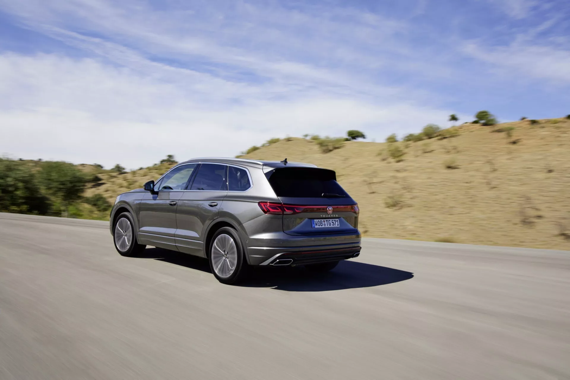 VW Touareg III lifting 2023 w ruchu na asfaltowej drodze, uchwycony z tylnego trzy-czwartego kąta ukazujący jego elegancki design, pięcioramienne koła ze stopu, metaliczne wykończenie, tylne światła LED, zintegrowane wydechy i sportową estetykę na tle suchej, pagórkowatej krajobrazu pod jasnym słonecznym niebem