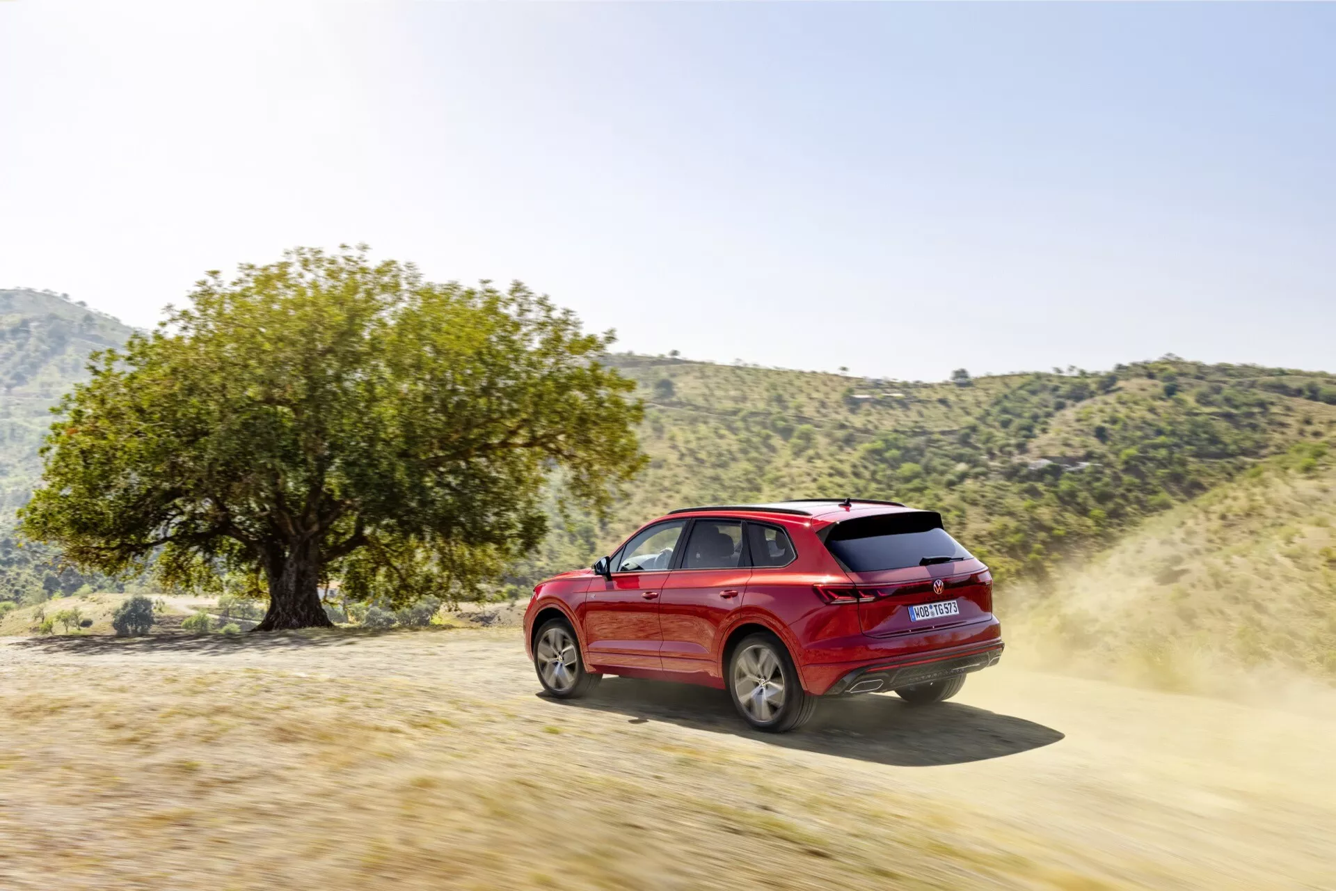 Czerwone SUV Volkswagen Touareg III Facelift 2023 demonstrujące zdolności terenowe na szutrowej drodze, widziane z trójkwartalnego tylnego kąta podkreślającego jego dynamiczny design, felgi, drzwi boczne, boczne i tylne okna, z kurzem wskazującym ruch i cechy wydajności takie jak konie mechaniczne i napęd 4x4, w naturalnym środowisku zewnętrznym sugerującym przygodę i wszechstronność terenową.