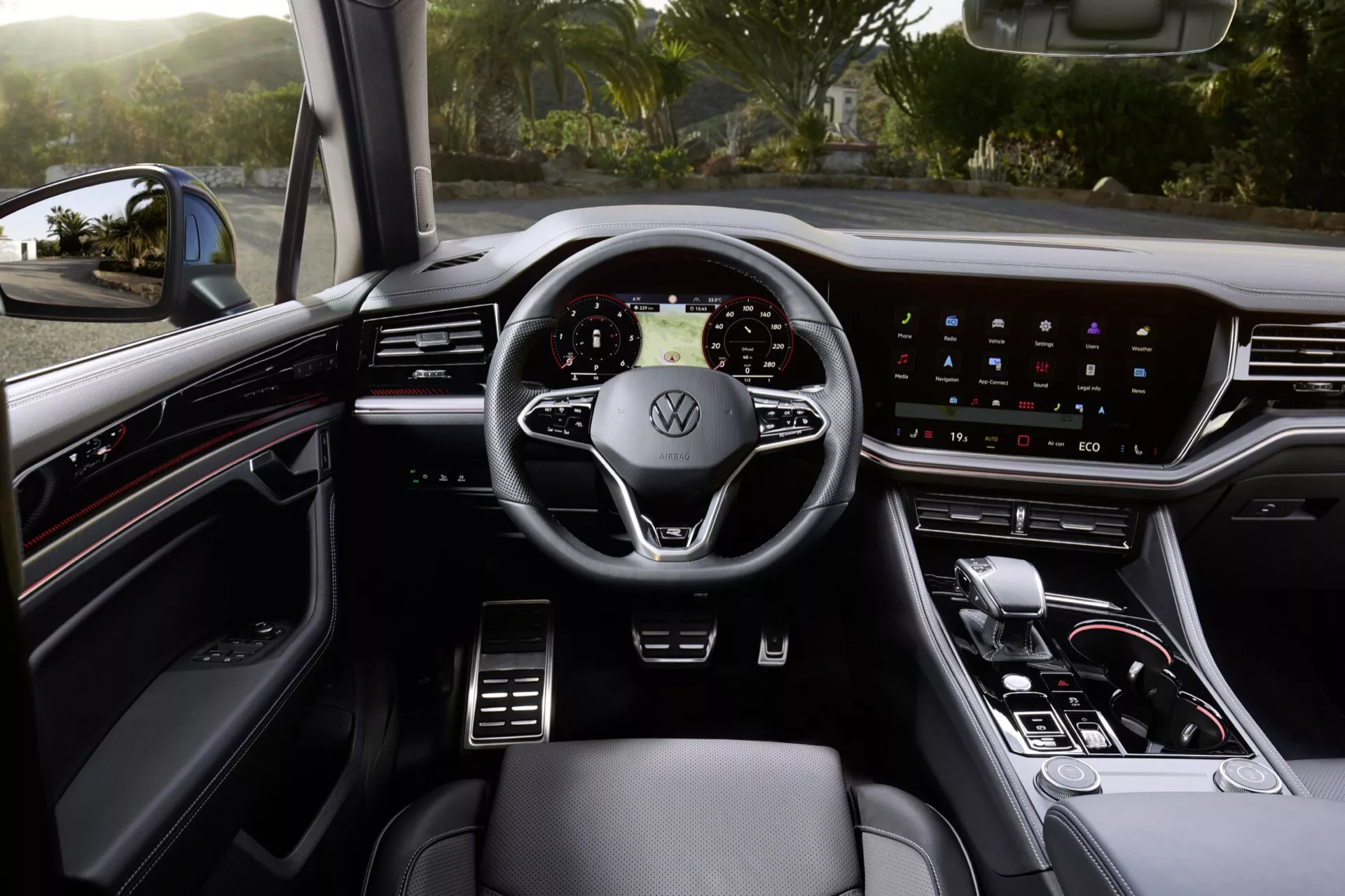 Wnętrze Volkswagen Touareg 2023 z perspektywy kierowcy pokazujące nowoczesną kierownicę trójramienną z logo Volkswagen, cyfrowy zestaw wskaźników, szeroki system infotainment z ekranem dotykowym na desce rozdzielczej, minimalne przełączniki fizyczne, dźwignię zmiany biegów automatycznych i premium skórzane siedzenia z widocznymi sterowaniami