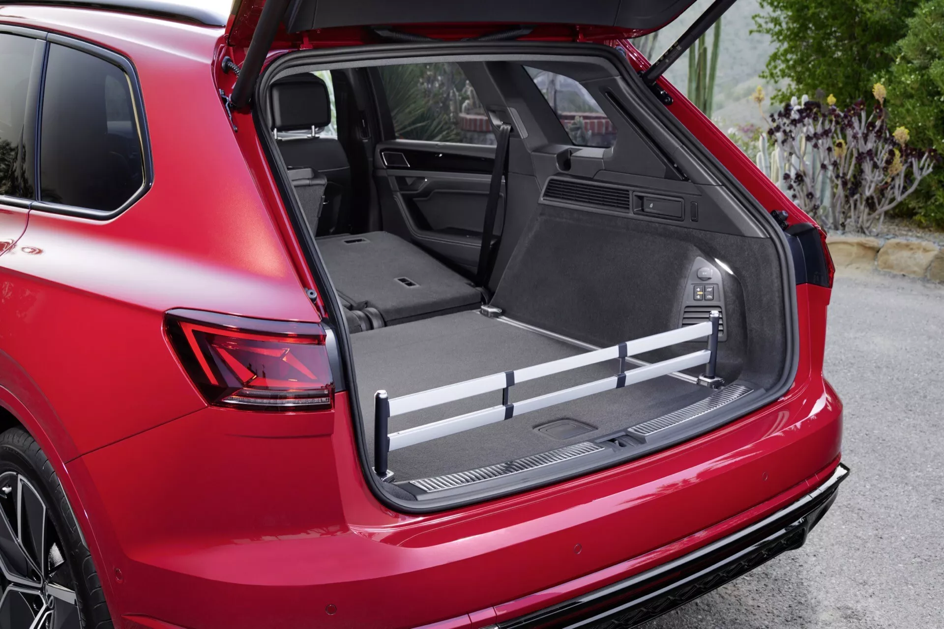 Zdjęcie szczegółowe Volkswagen Touareg III po liftingu 2023 pokazujące przestronny bagażnik z płaską podłogą załadunku, ochronną wykładziną i zintegrowanymi schowkami z widoku tylnego prawego pod kątem trzech czwartych, z wyróżniającym się gniazdem 12V, regulowanymi belkami bagażowymi i częściowo złożonymi tylnymi siedzeniami dla zwiększenia przestrzeni, podkreślone czerwonym kolorem nadwozia pojazdu i sportowym spojlerem.